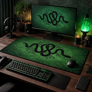 May include: A computer desk setup featuring a green and black mousepad with a snake design, a matching computer monitor, and a black keyboard. The desk also includes a black mouse, a smartphone, and a lava lamp.