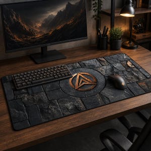 Può includere: Un tappetino per scrivania nero e grigio con una texture simile alla pietra e un emblema color bronzo. Una tastiera e un mouse neri sono sul tappetino, che si trova su una scrivania di legno. Un monitor di computer e una lampada da scrivania sono sullo sfondo.