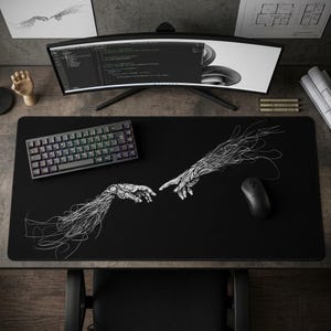 May include: Black rectangular desk mat featuring a white line art illustration of two hands almost touching, inspired by the Sistine Chapel. A black keyboard and mouse are on the mat. A curved monitor and other desk accessories are in the background.