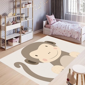 Può includere: Un tappetino da gioco beige con un'illustrazione di scimmia di cartone animato marrone. La scimmia ha un grande sorriso e guarda direttamente lo spettatore. Il tappetino è perfetto per la sala giochi o la camera da letto di un bambino.