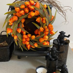 Può includere: Una ghirlanda circolare con vivaci fiori di alchechengi arancioni e lunghe foglie verdi sottili. La ghirlanda è esposta insieme a tre statuette di scimmie nere, un portacandele nero con una candela bianca e piccole zucche decorative.