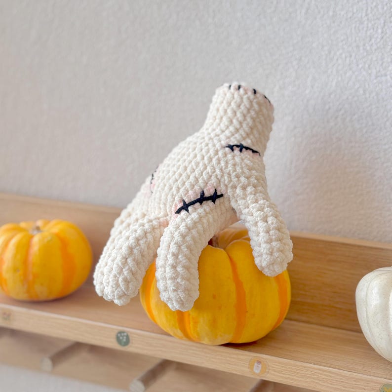 May include: A white crocheted plush toy shaped like a hand with stitched details, sitting on top of a yellow pumpkin. The pumpkin is on a wooden shelf.