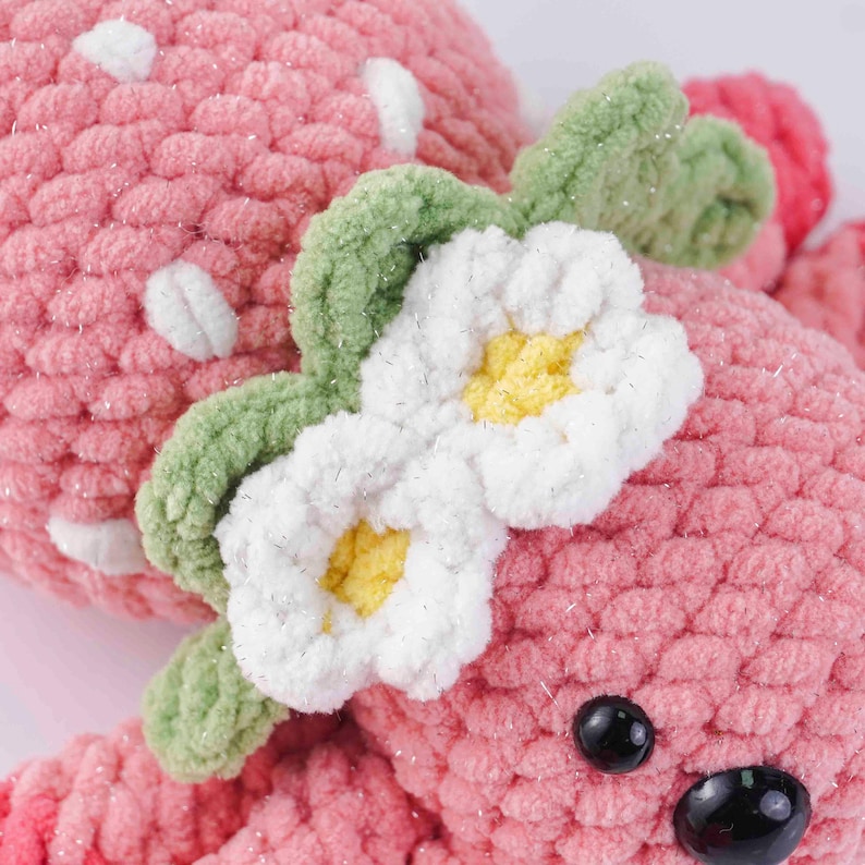 May include: A handmade pink crocheted strawberry plush toy with white spots, green leaves, and a white and yellow flower. The toy has black safety eyes and a sparkly texture.