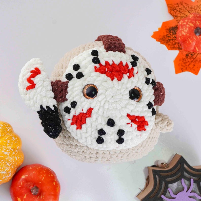 May include: A handmade crochet Jason Voorhees doll with a white mask featuring red and black details. The doll has brown accents and large, glittery eyes. It holds a white and red crochet knife. A Halloween-themed decorative item.