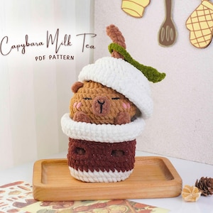 May include: A crocheted capybara milk tea plushie. The plushie is brown with a white hat and a maroon cup. The text "Capybara Milk Tea PDF Pattern" is above the plushie. The plushie is on a wooden tray.