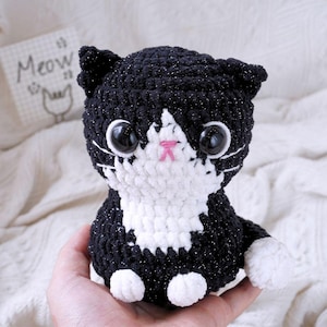 May include: Black and white crocheted cat plush toy with sparkly black fur and large black eyes. The cat is sitting with its paws tucked underneath it.