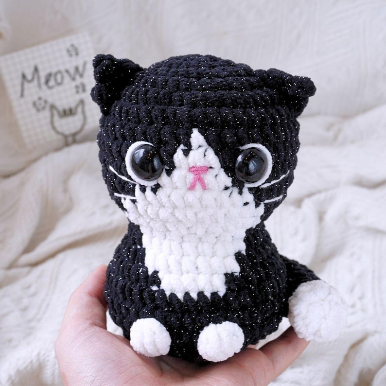 May include: Black and white crocheted cat plush toy with sparkly black fur and large black eyes. The cat is sitting with its paws tucked underneath it.