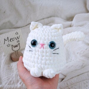 May include: White crocheted cat plush toy with blue eyes and a pink nose. The toy is sitting in a hand and has a long tail.
