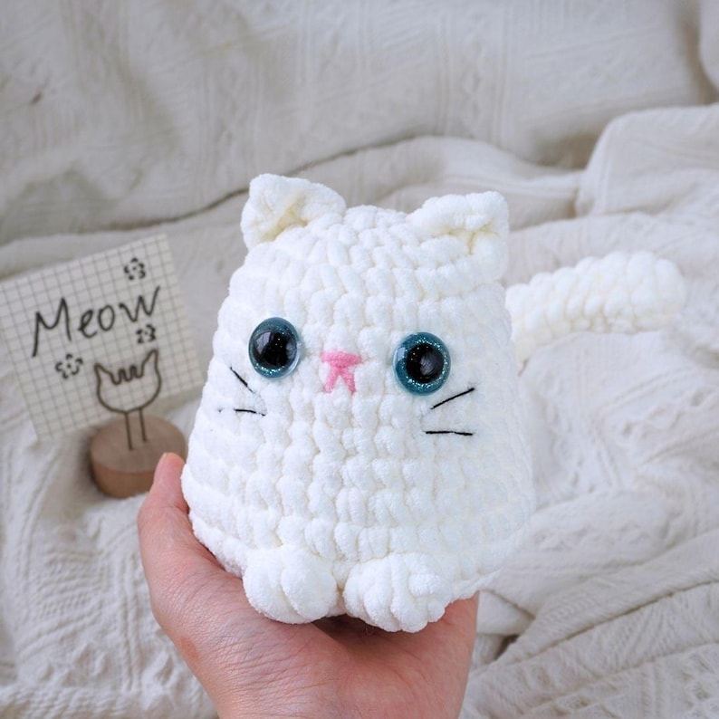 May include: White crocheted cat plush toy with blue eyes and a pink nose. The toy is sitting in a hand and has a long tail.
