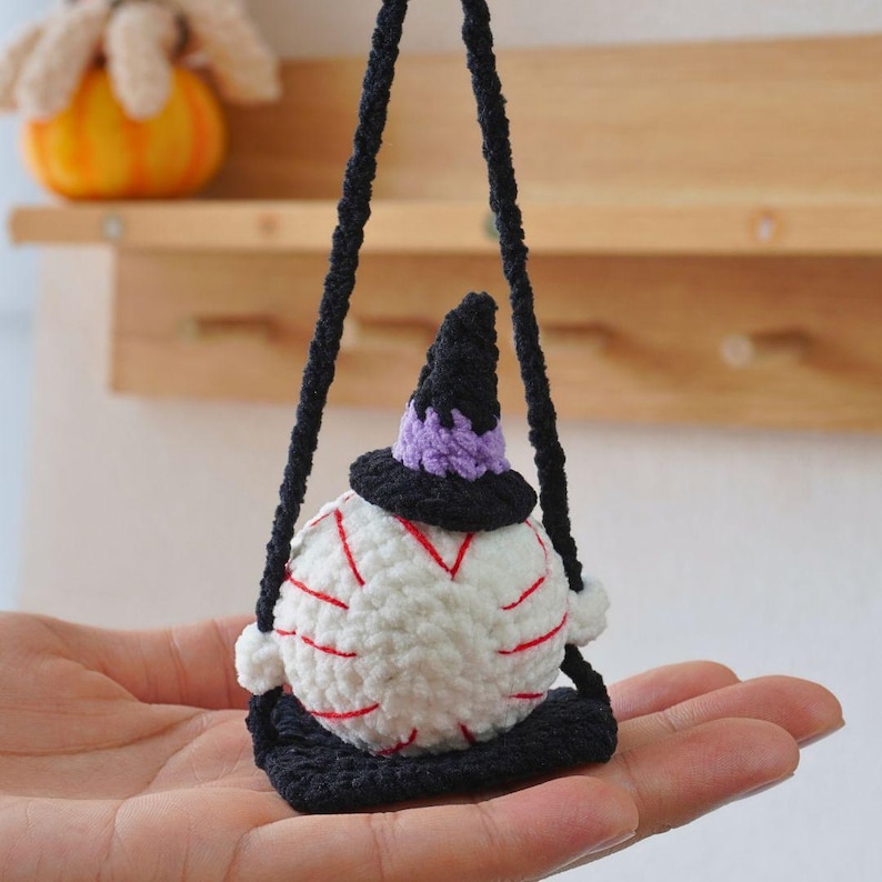 May include: A white crocheted ball with red stripes and a black witch hat is hanging from a black string. The ball is sitting on a black crocheted seat.