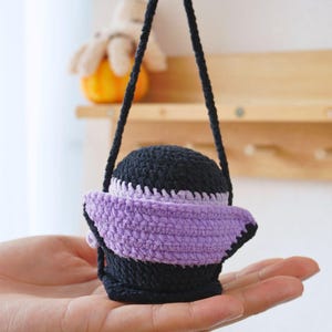 May include: A small crocheted bag with a black and purple design. The bag has a purple strap and is hanging from a hand.