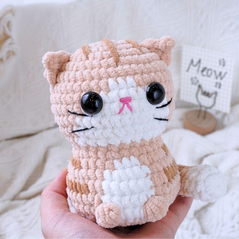 May include: A crocheted plush toy cat with orange and white fur. The cat has large black eyes, a pink nose, and a white face. The cat is sitting on a hand.