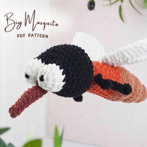 Pode incluir: Um brinquedo de mosquito feito à mão em crochê, com cabeça preta, olhos brancos e uma probóscide marrom. O corpo é laranja e marrom com asas brancas. O texto "Big Mosquito PDF Pattern" está acima.