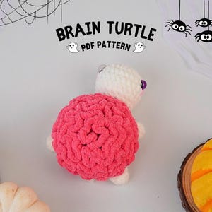 May include: A handmade crochet turtle with a pink brain-shaped shell and a white body. The text "BRAIN TURTLE PDF PATTERN" is displayed above the turtle. Halloween-themed decorations, including spiders and pumpkins, are in the background.