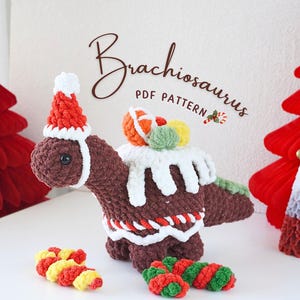 Puede incluir: Un Brachiosaurus de jengibre tejido a crochet con un gorro de Papá Noel y llevando adornos coloridos. El dinosaurio está decorado con glaseado blanco y bastones de caramelo rojos y blancos. La imagen incluye el texto "Brachiosaurus PDF PATTERN".
