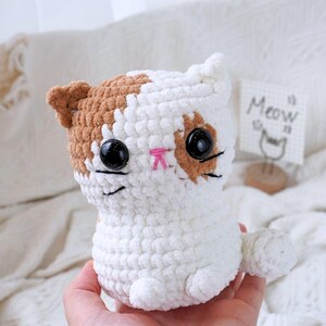 May include: A white and brown crocheted cat plush toy with black eyes and a pink nose. The toy is sitting on a person's hand.