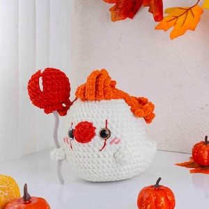 May include: A handmade crochet clown doll with a red nose, orange hair, and a red balloon. The doll is white with black eyes and pink cheeks. The clown is holding a red balloon with a gray stem. The doll is a Halloween or fall-themed item.