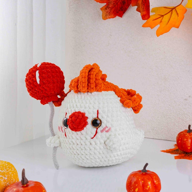 May include: A handmade crochet clown doll with a red nose, orange hair, and a red balloon. The doll is white with black eyes and pink cheeks. The clown is holding a red balloon with a gray stem. The doll is a Halloween or fall-themed item.