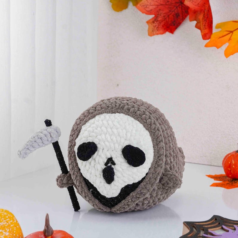 May include: A handmade, crocheted Halloween decoration featuring a round, gray figure with a white skull face and black eye sockets. It holds a small, gray scythe. The spooky plushie is a unique, festive item.