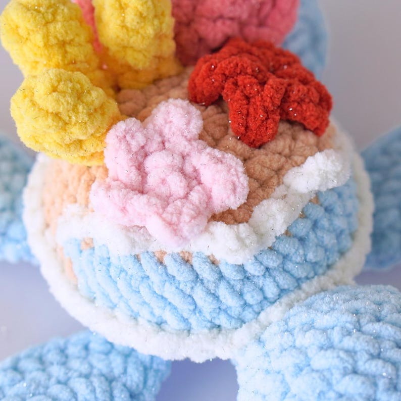 May include: A crocheted toy with a blue body, white trim, and a multi-colored top. The top features pink, yellow, and red crocheted flowers.