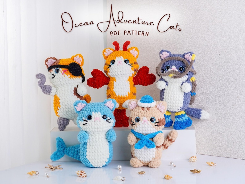 May include: A collection of five crocheted cat figures, each with a unique ocean-themed design. One cat wears an eye patch, another has a mermaid tail, and another wears a snorkel. The text "Ocean Adventure Cats PDF Pattern" is displayed above.