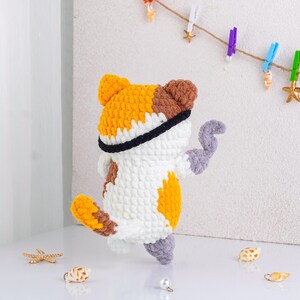 May include: A handmade crochet cat doll in white, orange, brown, and gray. The cat has a black band around its head and is posed with one arm raised. Decorative seashells and a pearl are scattered around the base.