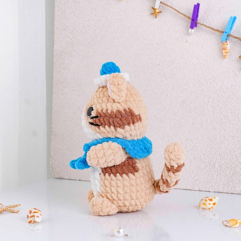 May include: A handmade crochet cat doll in tan, brown, and white, wearing a blue scarf and hat. The plush toy has black embroidered eyes and a striped tail. The cat is sitting on a white surface.