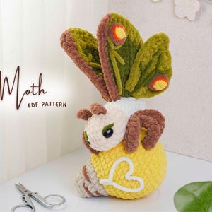 May include: A handmade crochet moth toy with large, detailed wings in shades of green, brown, and yellow. The moth's body is yellow with a white heart-shaped design. The text "Moth PDF Pattern" is visible.