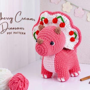 May include: A pink crocheted triceratops dinosaur with a white and red cherry-themed frill. The dinosaur has brown horns and black eyes. The text "Cherry Cream Dinosaur PDF Pattern" is in the upper left corner.