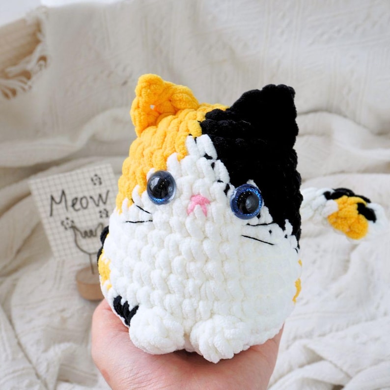 May include: A crocheted stuffed cat toy with white, black, and yellow yarn. The cat has blue eyes and a pink nose.