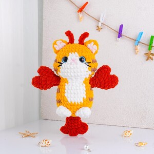 May include: A handmade crochet tiger toy with orange, white, and brown stripes. The tiger has red heart-shaped arms and feet, blue eyes, and a pink nose. The toy is standing on a white surface.