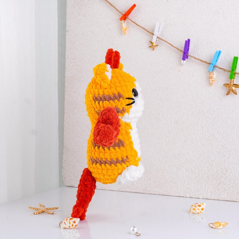 May include: A handmade crochet cat doll in orange and brown stripes, with white accents and a red tail. The doll has black eyes and a red tuft of hair. The background includes a string with decorative clothespins and seashells.