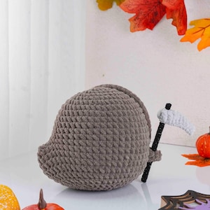 May include: A handmade, gray crocheted Grim Reaper figure with a black and white scythe. The plushie is round and has a small, pointed head. The background includes fall leaves and pumpkins, suggesting a Halloween theme.