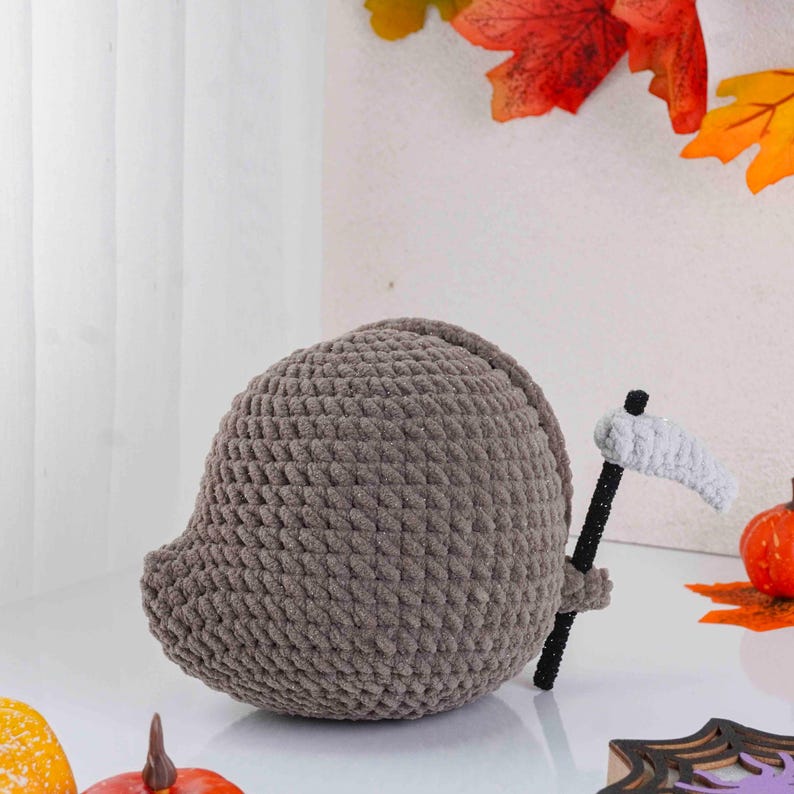 May include: A handmade, gray crocheted Grim Reaper figure with a black and white scythe. The plushie is round and has a small, pointed head. The background includes fall leaves and pumpkins, suggesting a Halloween theme.