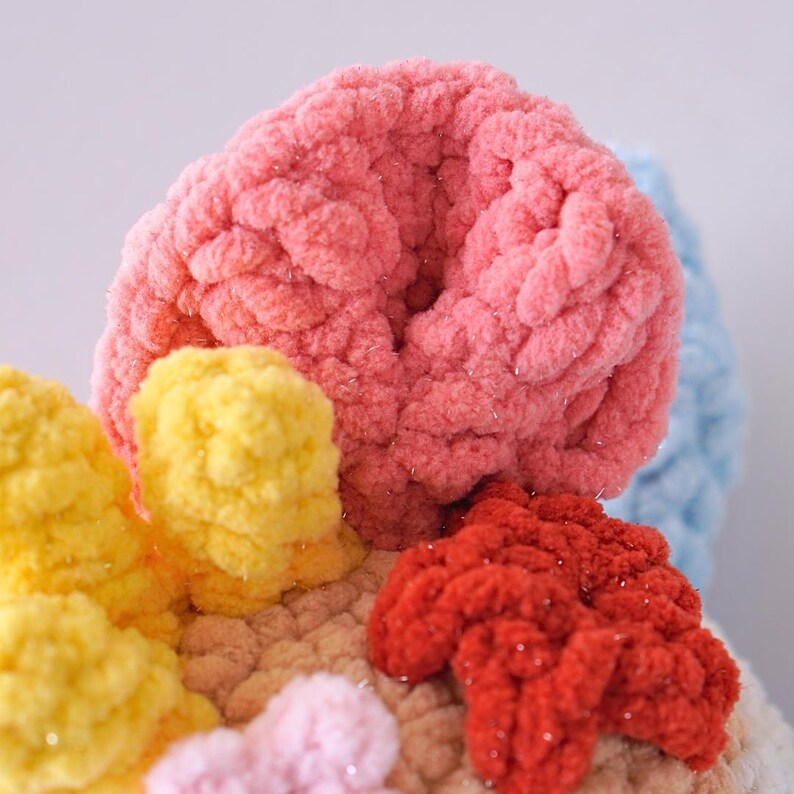 May include: Close-up of a crocheted coral reef sculpture with pink, yellow, red, and blue yarn. The coral is made with a textured, fluffy yarn.