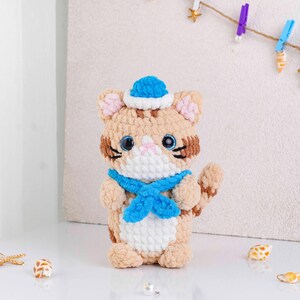 May include: A handmade crochet cat doll with a blue sailor hat and scarf. The cat has tan and white fur with brown stripes, large blue eyes, and a pink nose. The doll is standing upright.