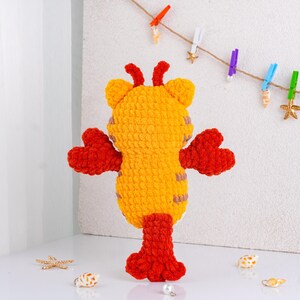 May include: A handmade, plush toy in the shape of a lobster. The toy is primarily yellow with red claws, legs, and antennae. The lobster has a textured, fuzzy appearance and is standing upright. Decorative seashells and a string with clothespins are in the background.
