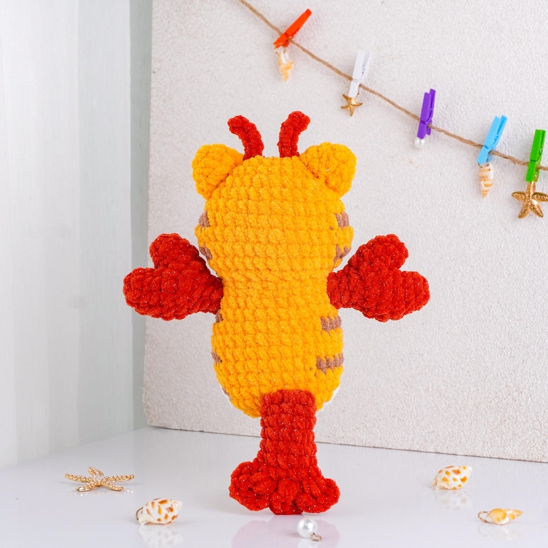 May include: A handmade, plush toy in the shape of a lobster. The toy is primarily yellow with red claws, legs, and antennae. The lobster has a textured, fuzzy appearance and is standing upright. Decorative seashells and a string with clothespins are in the background.