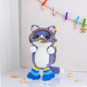 May include: A handmade crochet cat doll in shades of blue, white, and yellow, wearing a diving mask and flippers. The cat has blue eyes and pink inner ears. Decorative seashells and starfish are scattered around the doll.