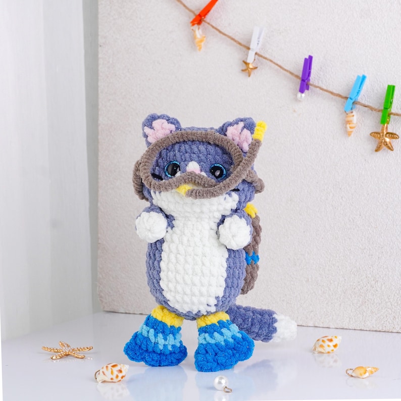 May include: A handmade crochet cat doll in shades of blue, white, and yellow, wearing a diving mask and flippers. The cat has blue eyes and pink inner ears. Decorative seashells and starfish are scattered around the doll.