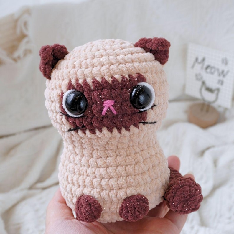 May include: A crocheted plush toy cat with brown and beige fur, large black eyes, and a pink nose. The cat is sitting on a hand.