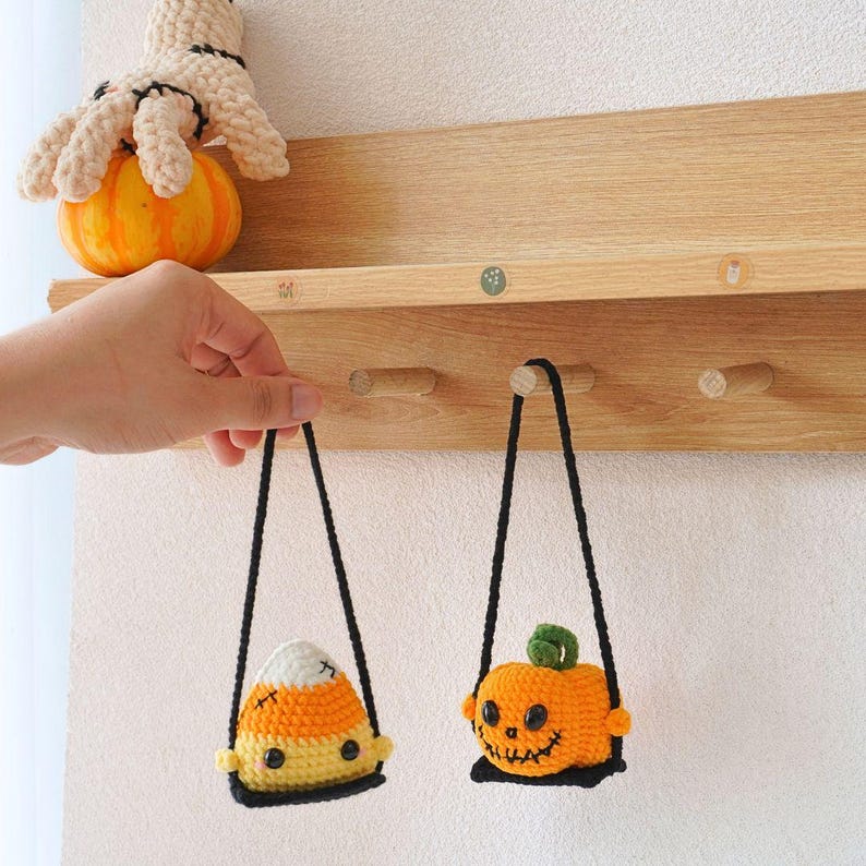 May include: Two crocheted Halloween decorations hanging from hooks on a wooden shelf. One is a candy corn and the other is a pumpkin with a stitched-on face.