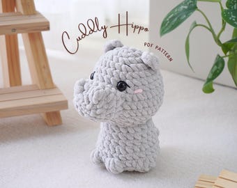 May include: A gray crocheted hippopotamus amigurumi toy with black eyes and pink blush. The toy is sitting on a white surface. The text "Cuddly Hippo PDF Pattern" is visible in the background.