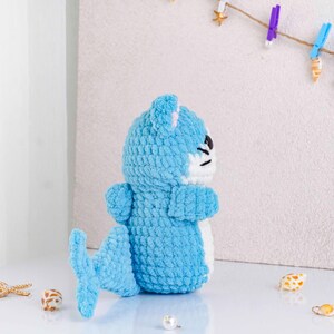 May include: A handmade blue crochet seal plush toy with a white belly and a small tail. The seal has black eyes and a white muzzle. The plush is sitting on a white surface. Decorative items are in the background.