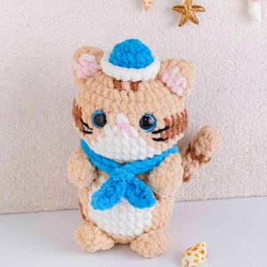 May include: A handmade plush cat with a blue sailor hat and scarf. The cat is tan with brown stripes, pink inner ears, and large blue eyes. The scarf and hat are blue with white trim. The cat is a cute, nautical-themed stuffed animal.