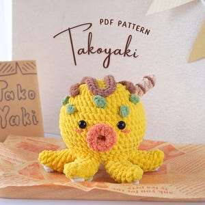 May include: A crocheted octopus plush toy shaped like a takoyaki, a Japanese street food. The octopus is yellow with brown and green accents. The toy has a pink and white face with black eyes and a pink donut-shaped mouth.