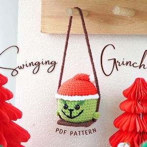Puede incluir: Un muñeco de Grinch de crochet con un gorro de Papá Noel rojo, colgado de una cuerda de columpio marrón. El muñeco es verde con un borde blanco y tiene una expresión traviesa. El fondo presenta árboles de Navidad de papel rojo y verde y el texto "swinging Grinch" y "PDF PATTERN".