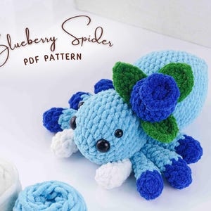 Könnte beinhalten: Hellblaue gehäkelte Spinne mit dunkelblauen Akzenten und einer Blaubeerblüte. Die Spinne hat schwarze Augen und weiße Beine. Der Text "Blueberry Spider PDF Pattern" ist in einer Schreibschrift geschrieben.