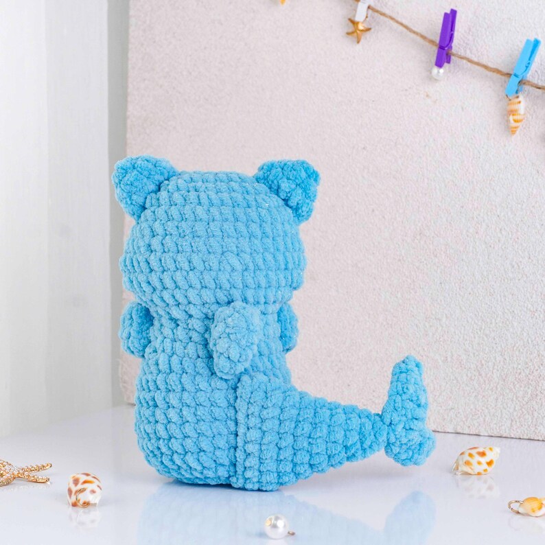 May include: A light blue, handmade crochet mermaid bear doll. The plush toy has a bear-like head with small ears, arms, and a mermaid tail. The doll is made of a soft, textured yarn. Decorative seashells and a pearl are in the foreground.