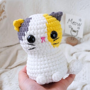 May include: A crocheted stuffed cat toy made with white, grey and yellow yarn. The cat has black eyes and a pink nose.